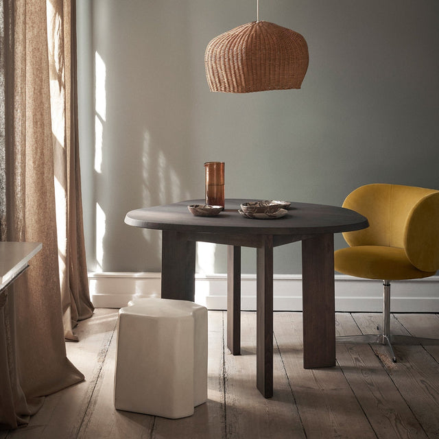 ferm LIVING Wohnzimmer – runder Esstisch aus dunklem Holz mit Rattan-Pendelleuchte und gelbem Sessel, skandinavische Möbel in warmem Raumlicht
